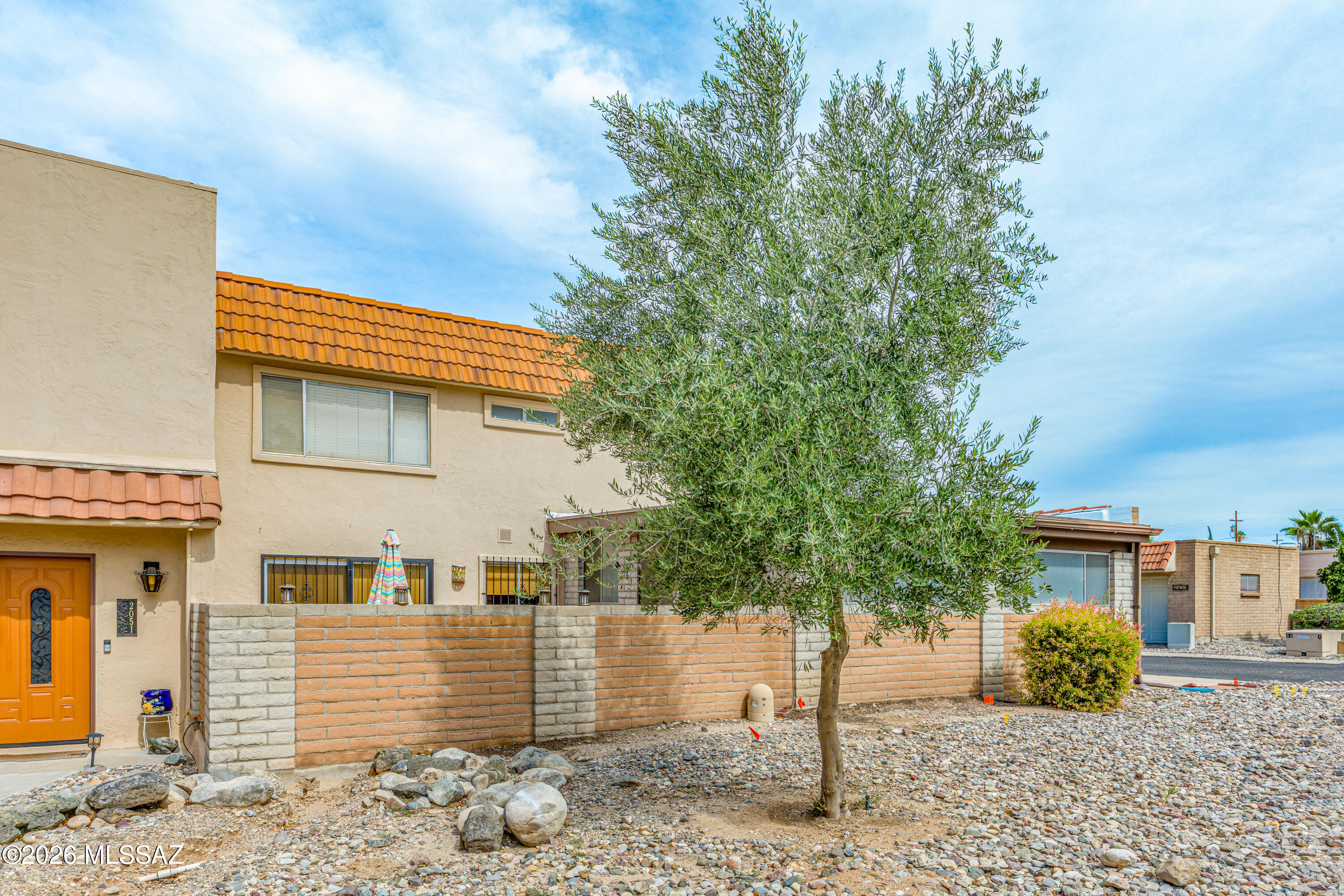 Beige stucco home with a terracotta tile roof, brick courtyard wall, and gravel yard beneath a mature tree.