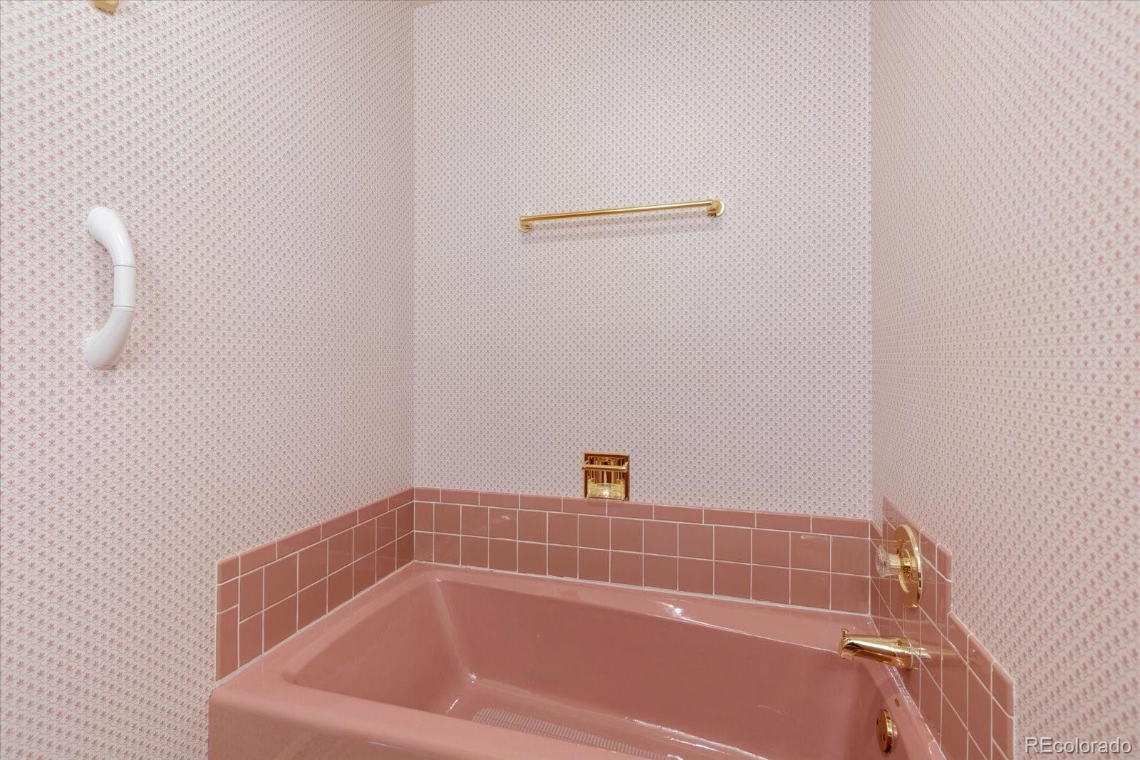 Pink bathtub with matching tile, patterned wallpaper, and gold-tone fixtures.