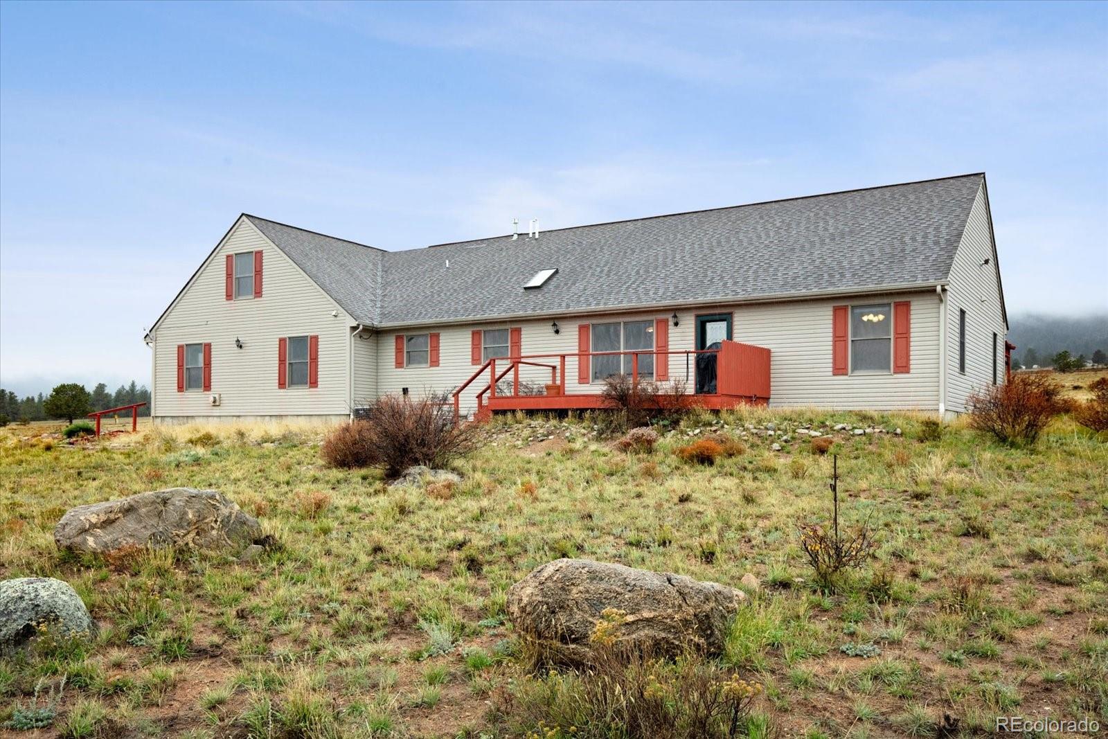 Light-colored house with red shutters and a red deck on rocky, grassy terrain.