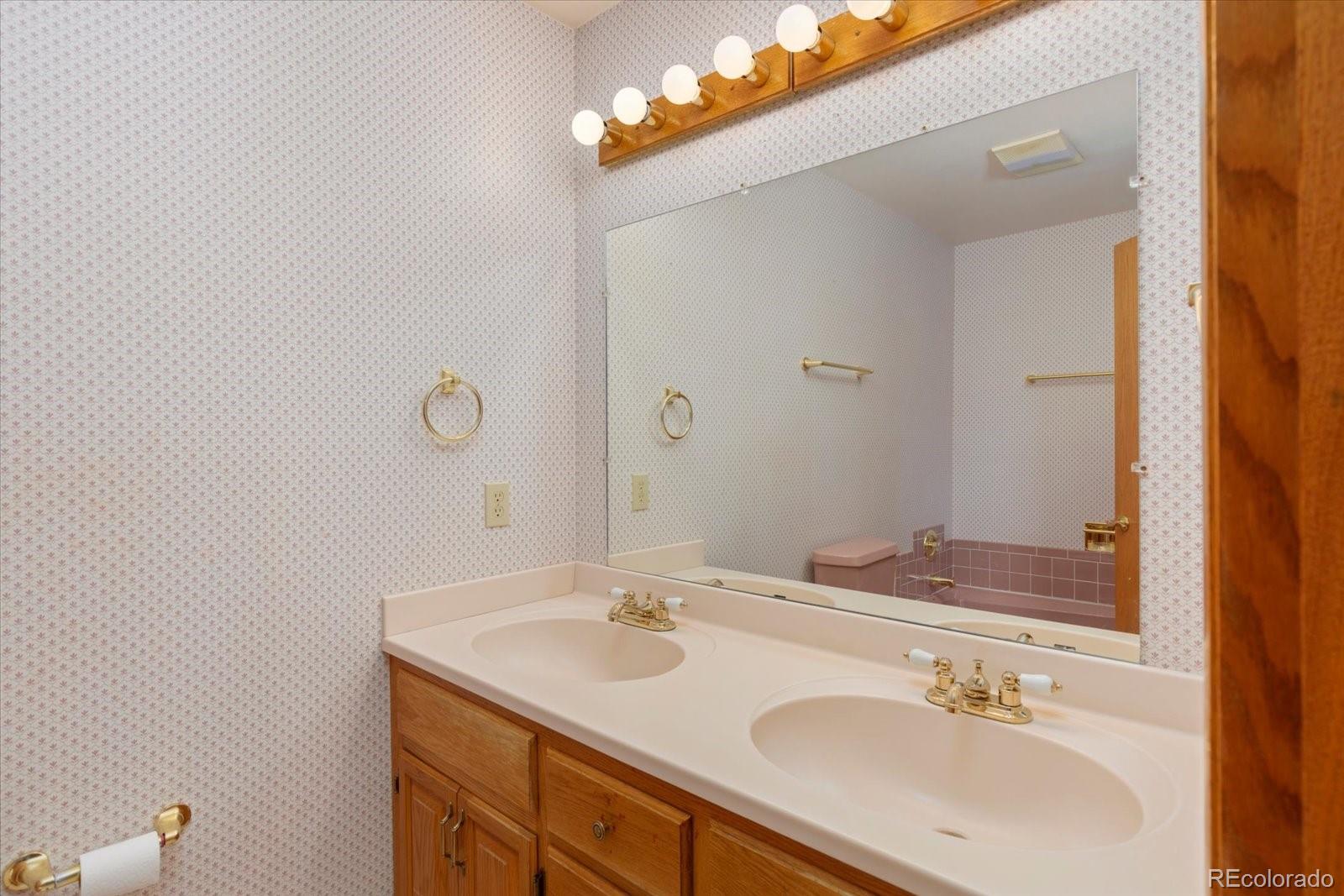 Bathroom with a double-sink wood vanity, large mirror, patterned wallpaper, and brass fixtures.