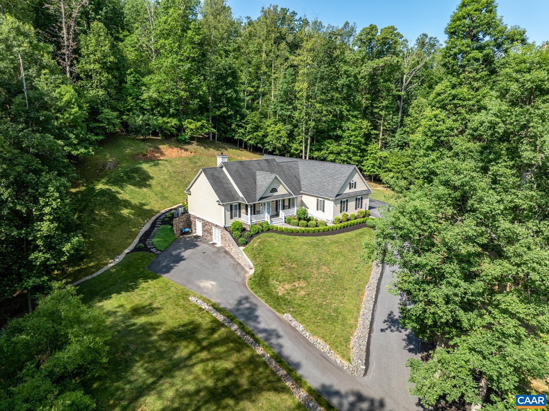 Aerial view of a house with a covered porch, landscaped yard, and curved driveway surrounded by trees.