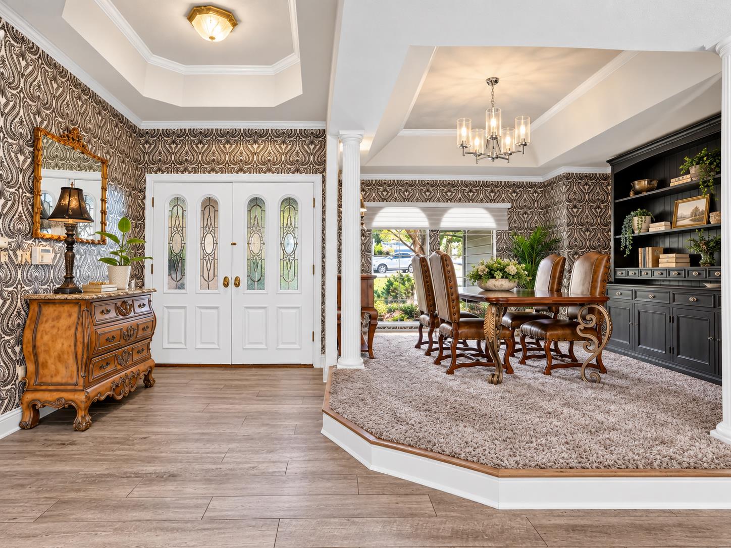 Formal dining room with a carved wood table, leather chairs, patterned wallpaper, and glass-paneled entry doors.
