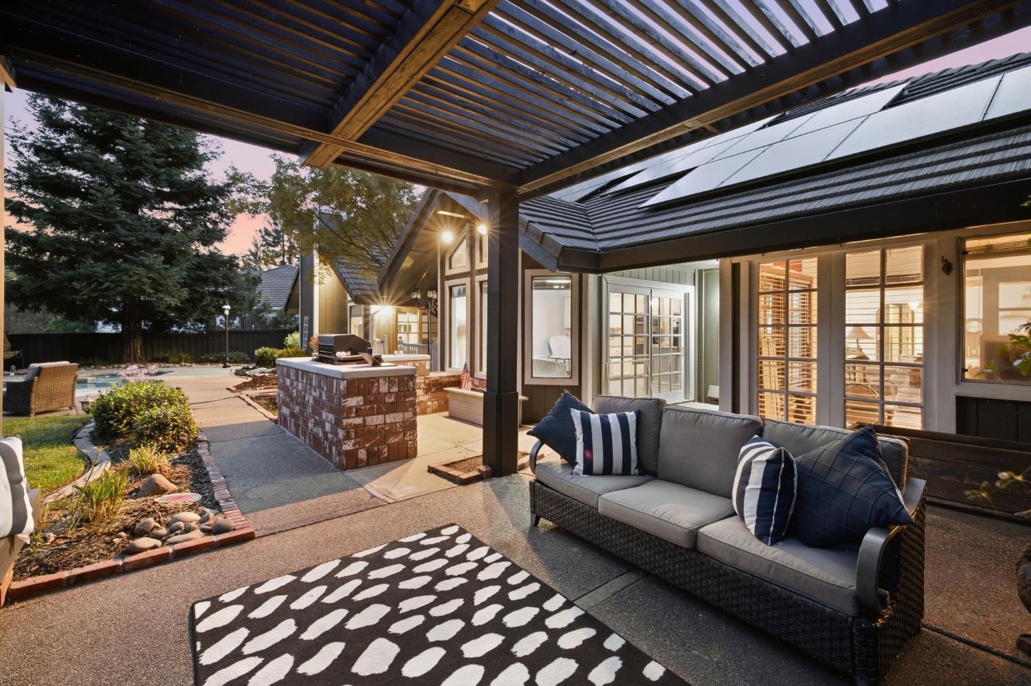 Covered patio with pergola, cushioned seating, brick grill island, and swimming pool at dusk.