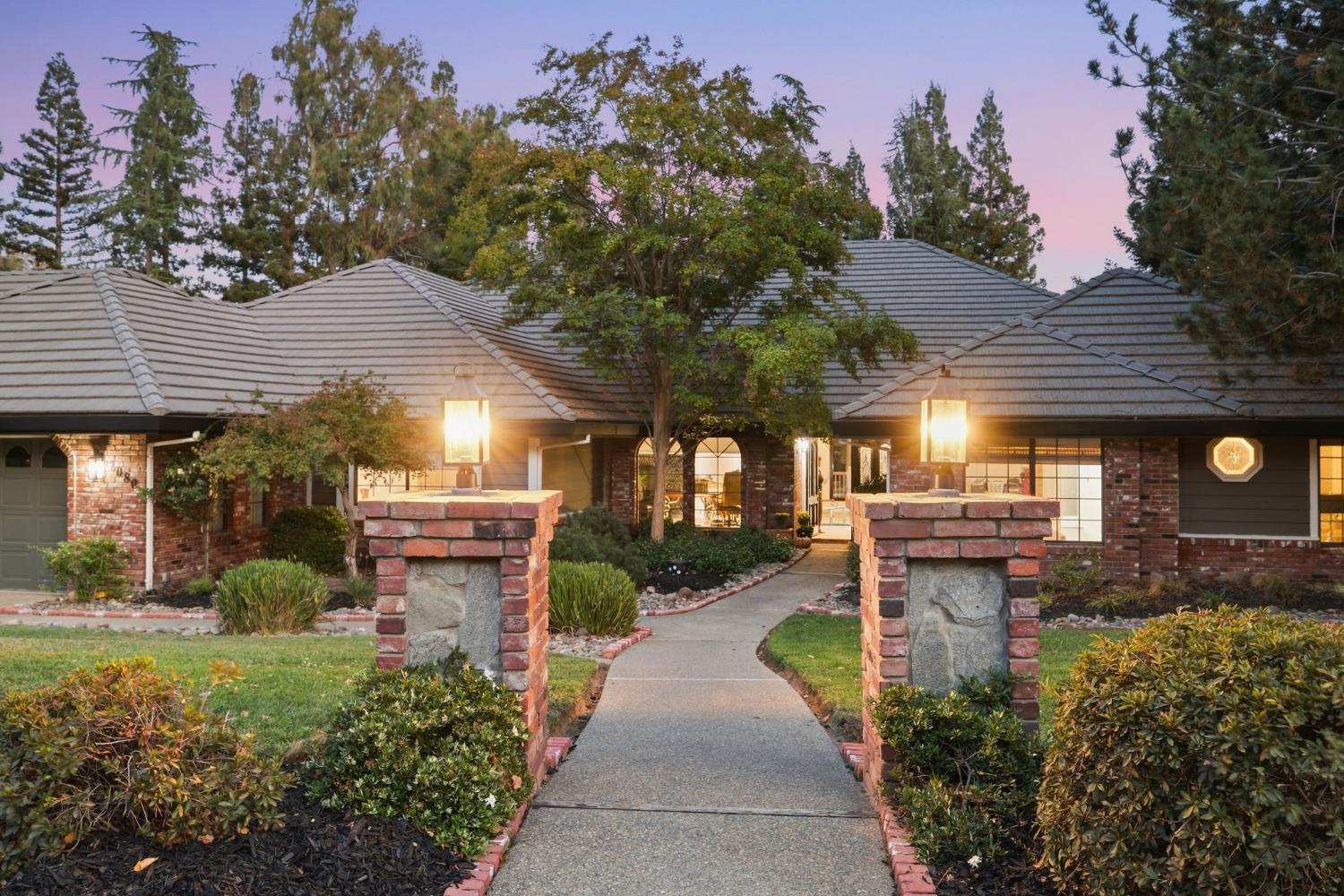 Single-story brick home at dusk, approached by a curved walkway between stone-and-brick pillars.
