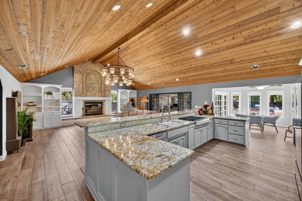 Open-plan kitchen with a large granite island, vaulted wood ceiling, and stone fireplace.