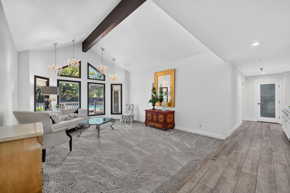 Living room with vaulted ceiling, dark beam, tall windows, and a glimpse of the pool.