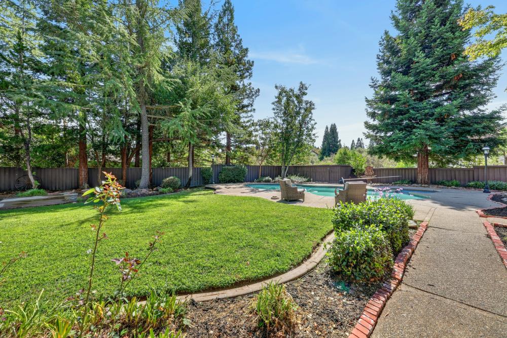 Backyard pool surrounded by mature trees, lawn, patio chairs, and a paved walkway.