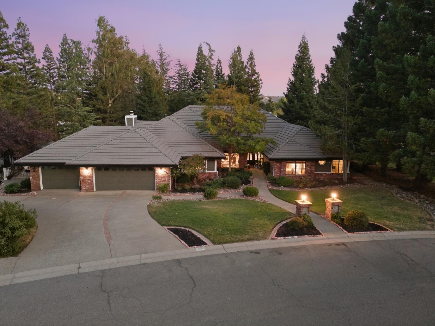Single-story brick home with a three-car garage, curved driveway, and illuminated entry pillars at dusk.