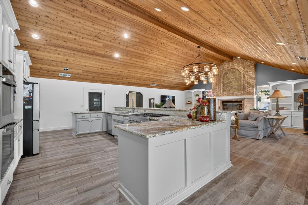 Kitchen with a large granite island and vaulted wood ceiling, opening to a living room with a brick fireplace.