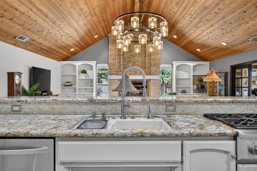 Kitchen with a granite island and sink beneath a vaulted wood ceiling, facing a brick fireplace.