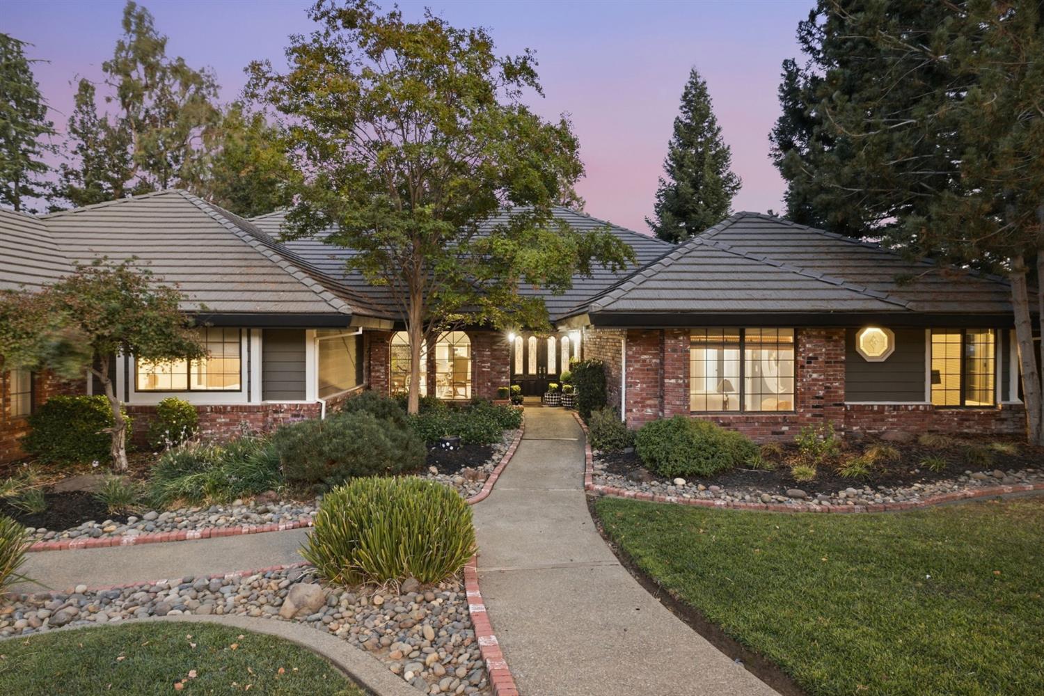 Single-story brick-and-gray home with a tiled roof, landscaped entry, and warmly lit windows at dusk.