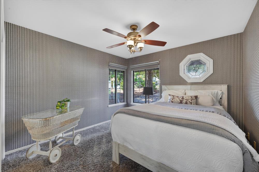 Bedroom with a white bed, patterned wallpaper, corner windows, and a wheeled wicker cart.