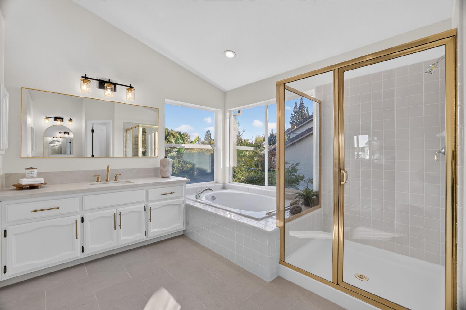 Bathroom with a tiled soaking tub beneath windows and a glass shower with gold trim.