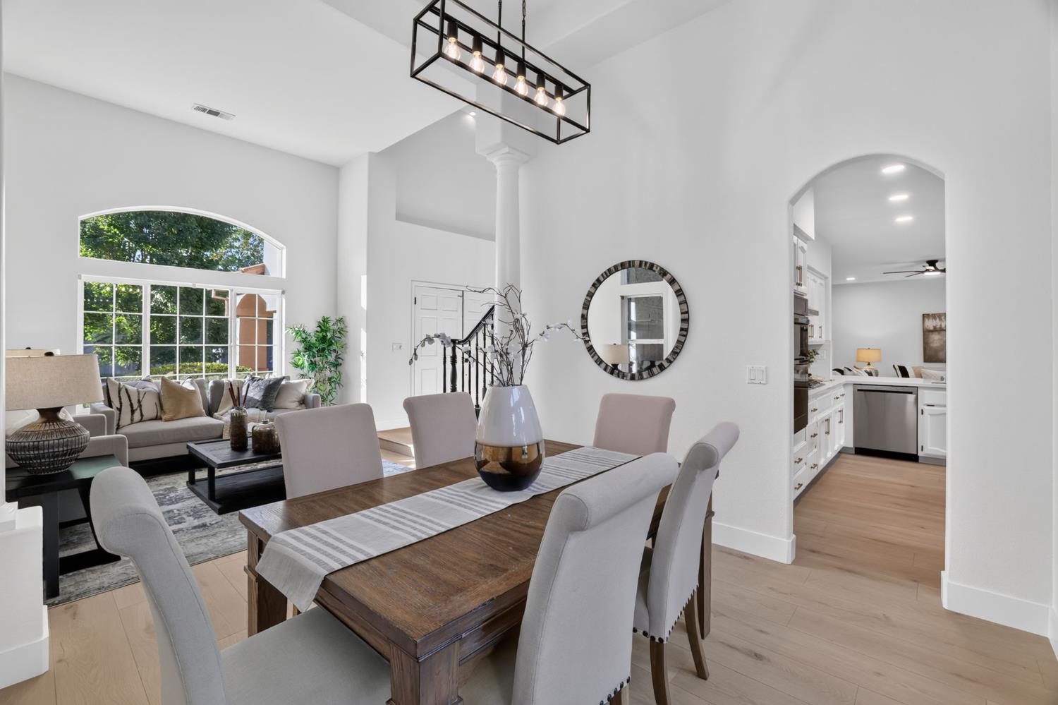 Open-plan dining and living room with a wood table, large arched window, and kitchen beyond.