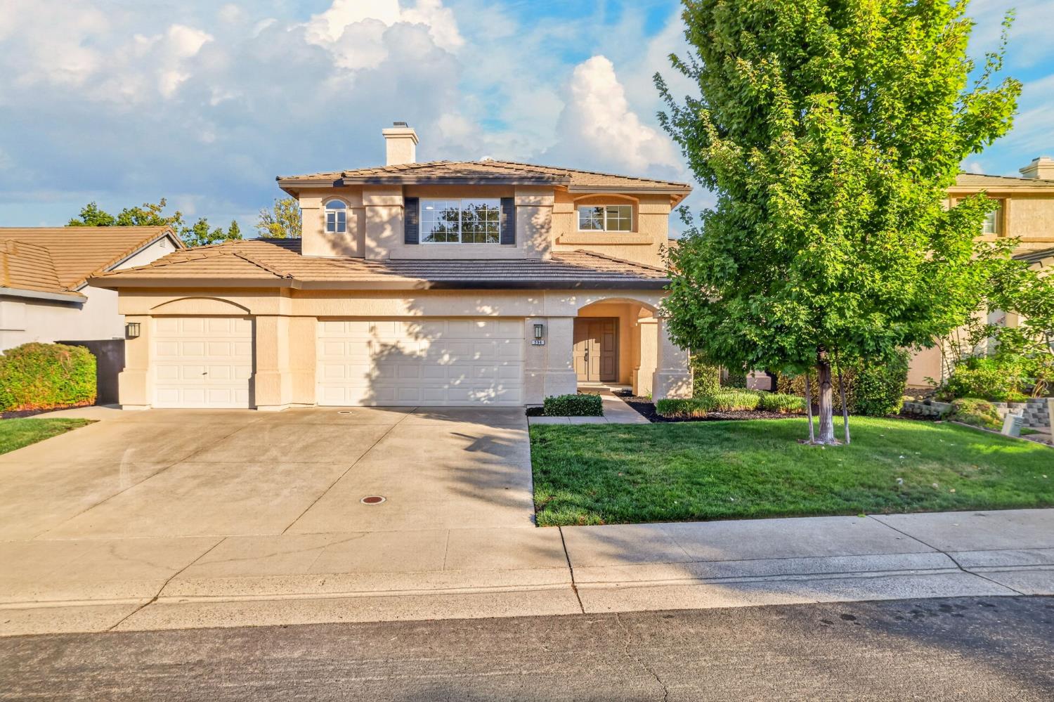 Two-story tan home with a three-car garage, arched entry, and large leafy tree in the front yard.