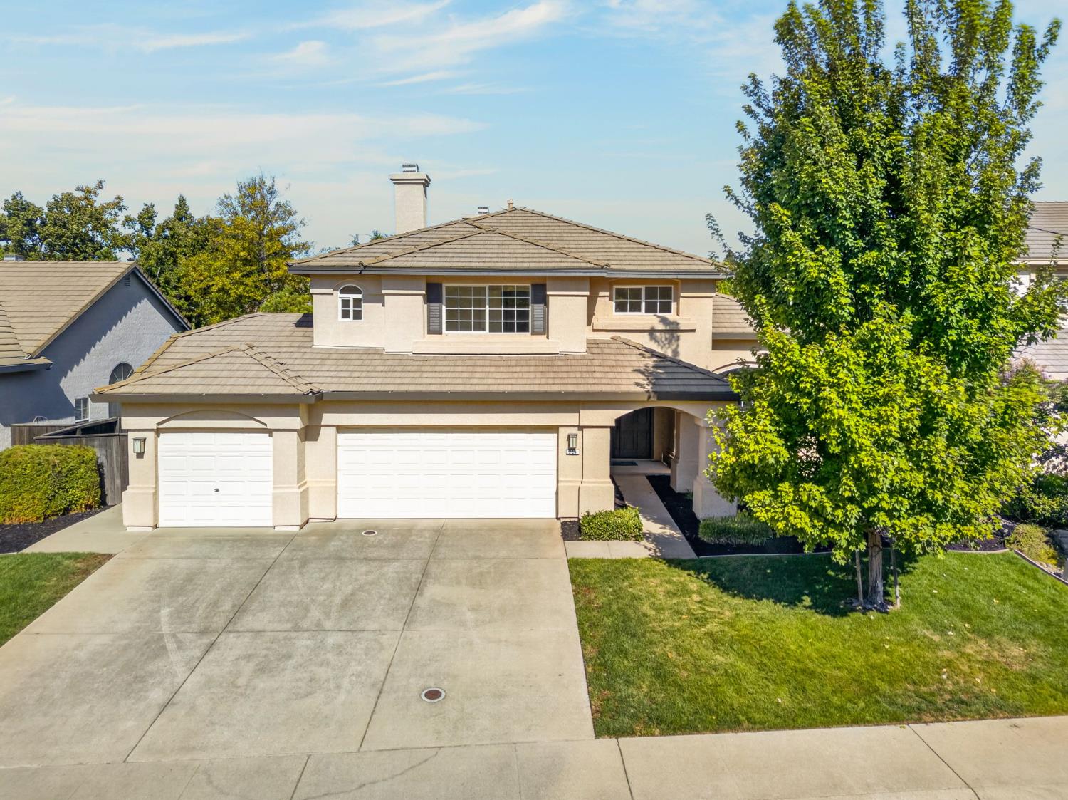 Two-story beige house with a tiled roof, double garage, wide driveway, and large tree in front.