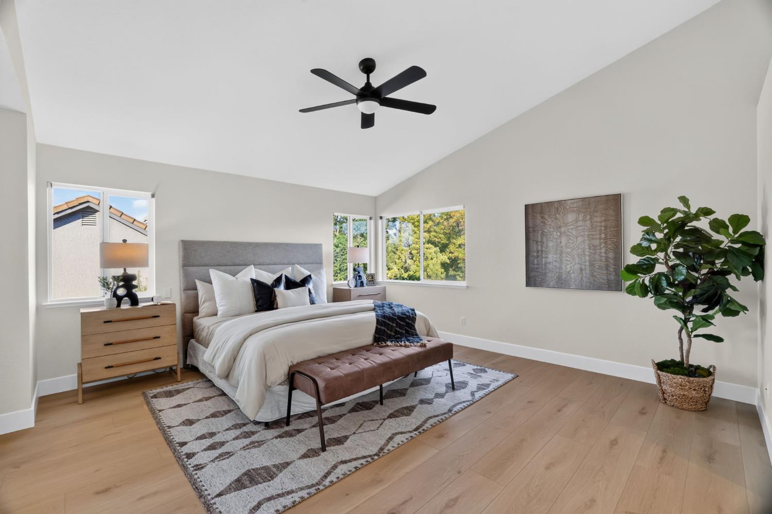 Staged primary bedroom with vaulted ceiling, king-size bed, light wood floors, and large windows.