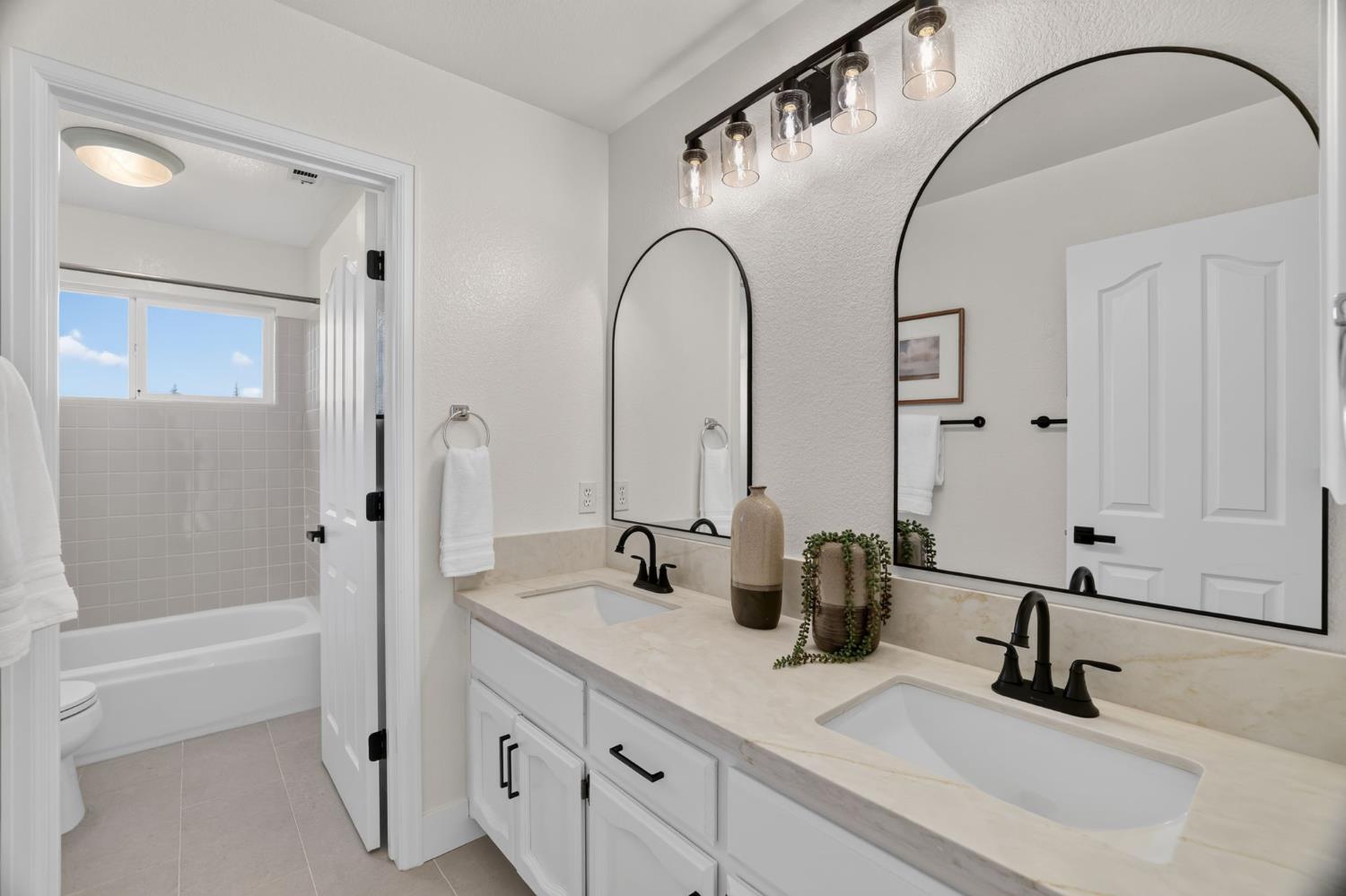 Bright bathroom with a double-sink vanity, arched mirrors, and an adjoining tub.