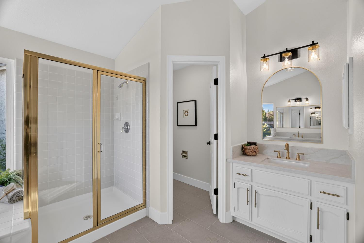 Bathroom with a gold-framed glass shower, white vanity, and arched mirror.