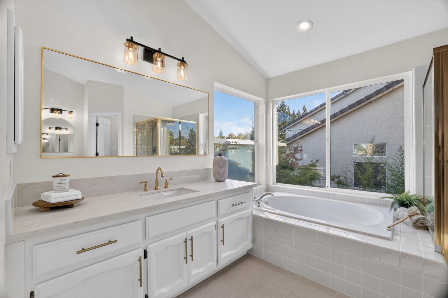 Bright bathroom with a white vanity, gold fixtures, and a built-in tub beside large windows.