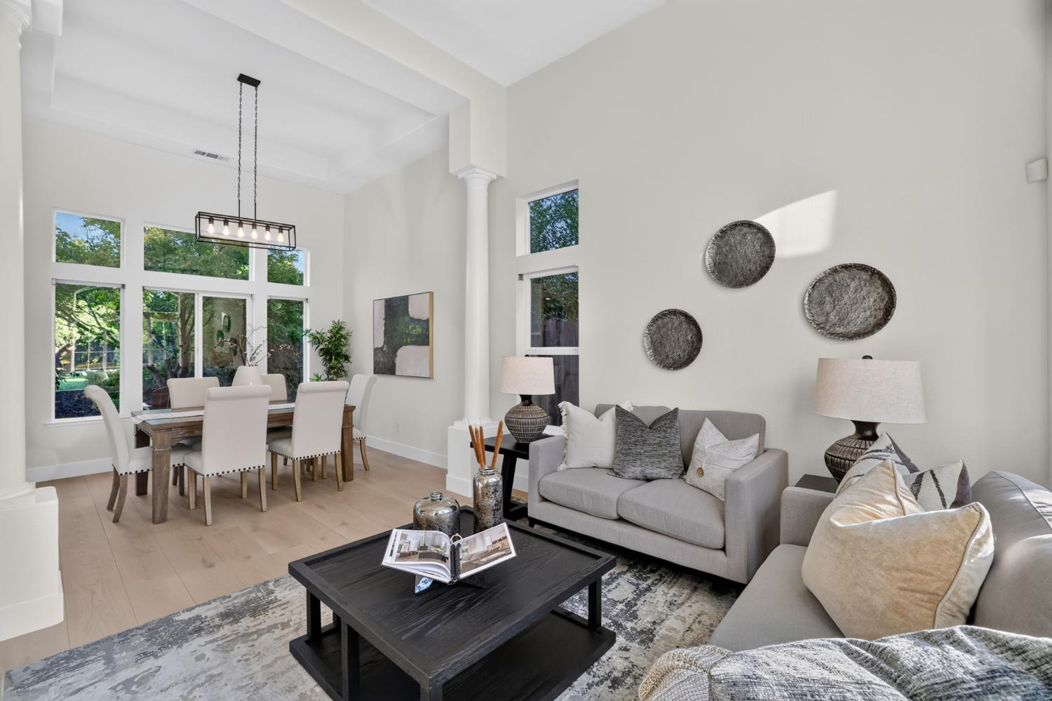 Staged living and dining room with vaulted ceiling, tall windows, gray sofas, and a black coffee table.