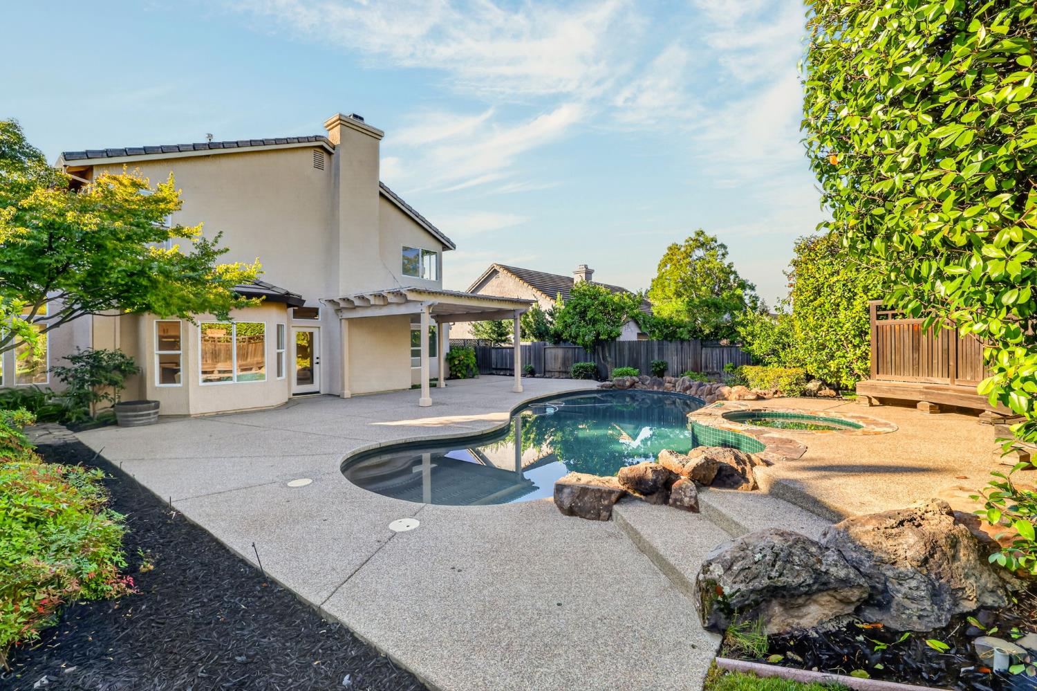 Backyard pool and spa beside a covered patio, surrounded by rock landscaping and mature greenery.