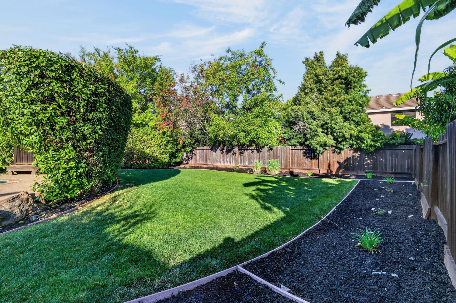 Backyard lawn bordered by mature trees, mulched garden beds, and a wooden privacy fence.