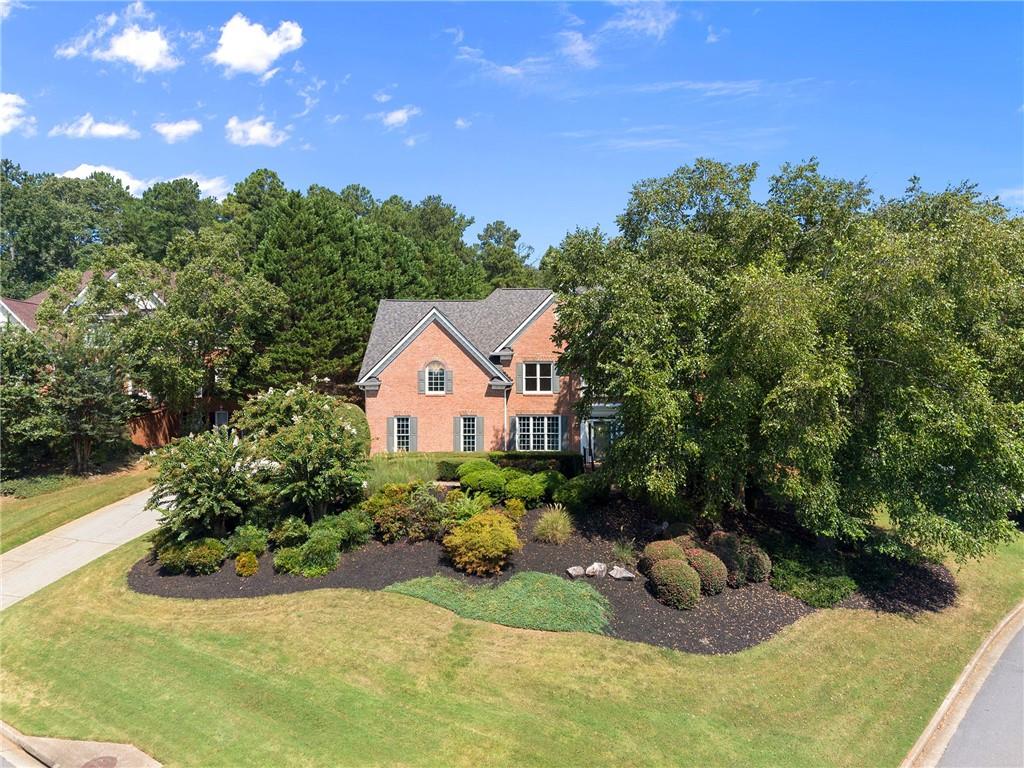Two-story red brick house with gray gabled roofs, mature trees, and landscaped garden beds.