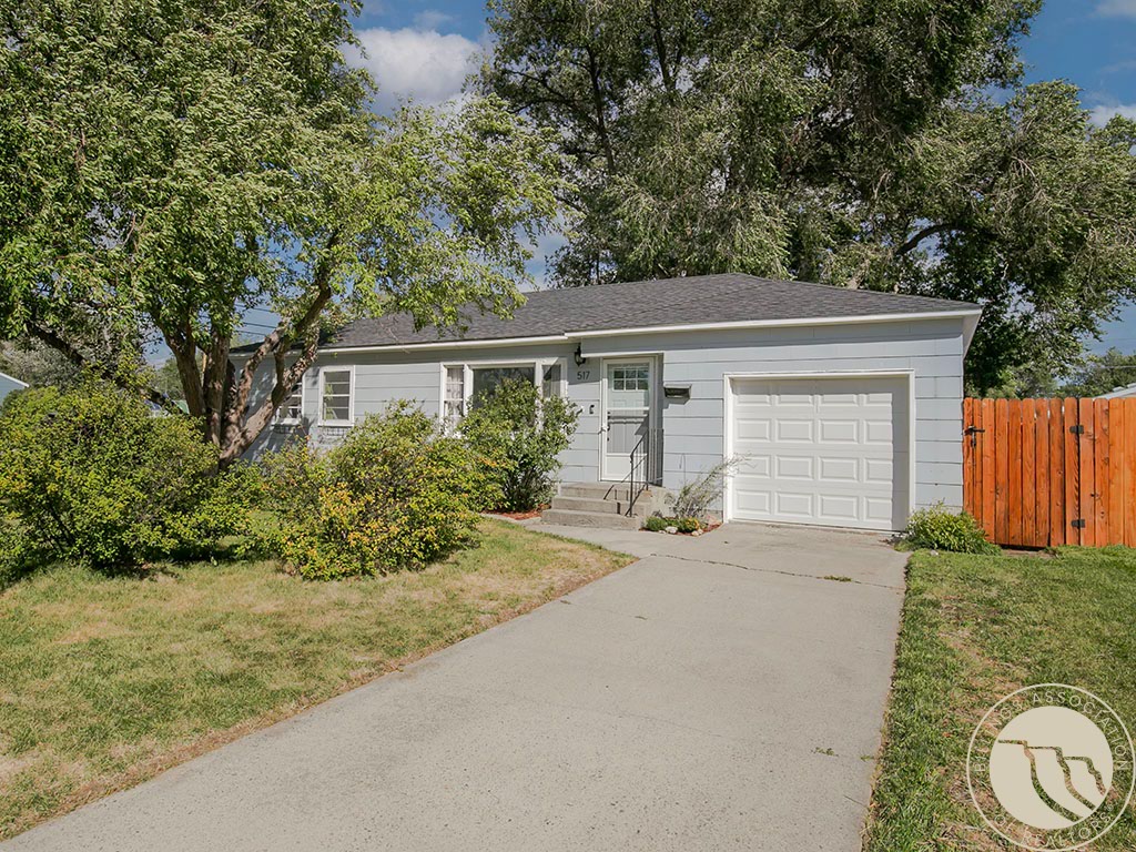 Light gray ranch-style house with an attached garage, mature trees, and a wooden fence.
