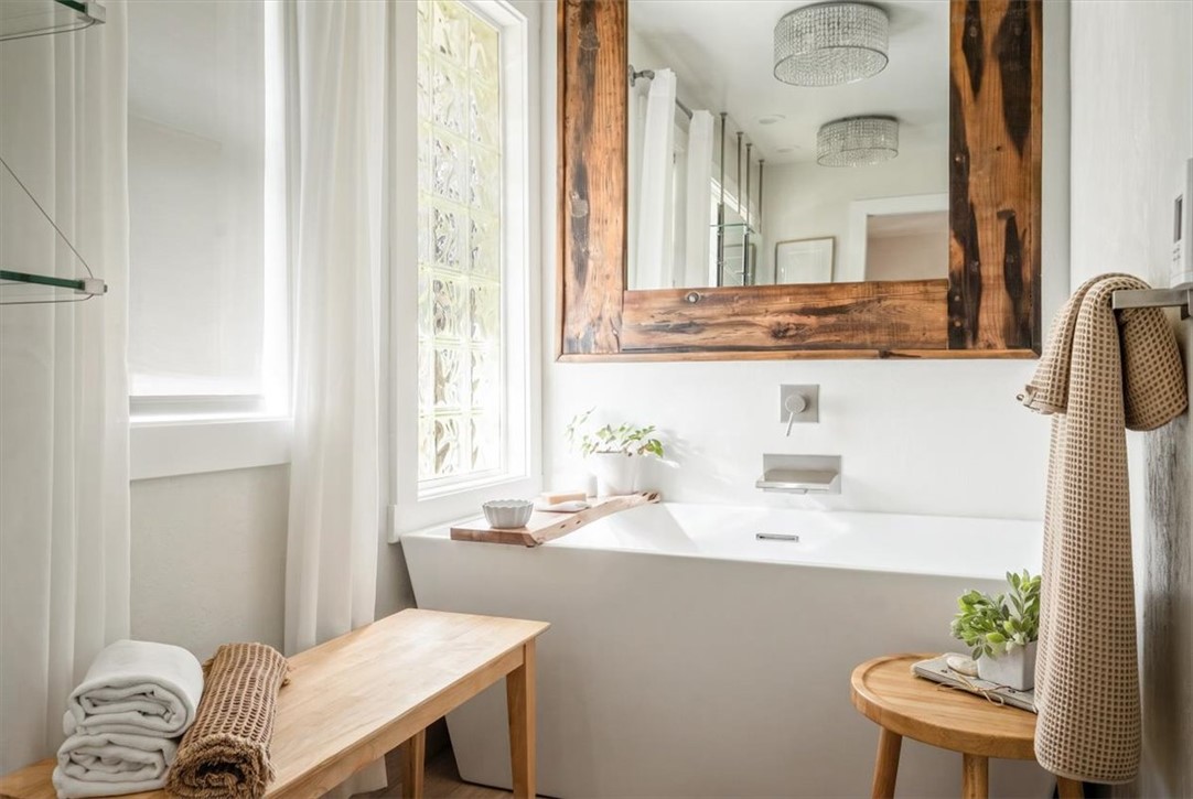 Bright bathroom with a white bathtub, rustic wood-framed mirror, tall windows, and wooden benches.