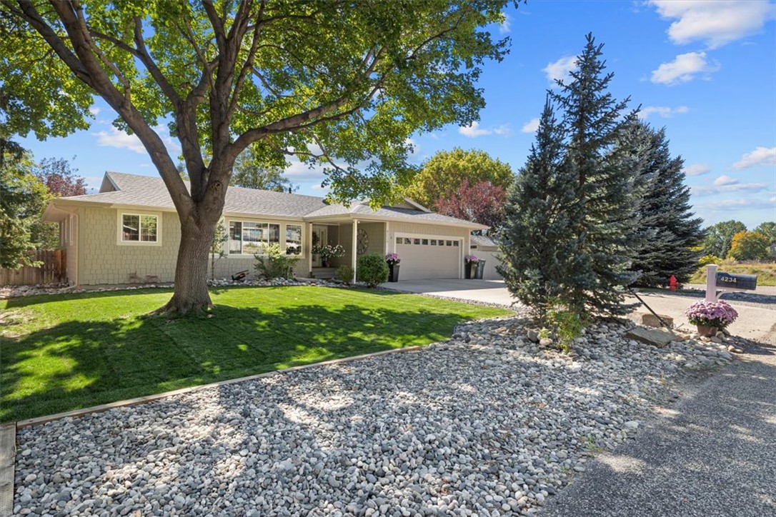 Single-story home with an attached garage, mature shade tree, green lawn, and rock landscaping.