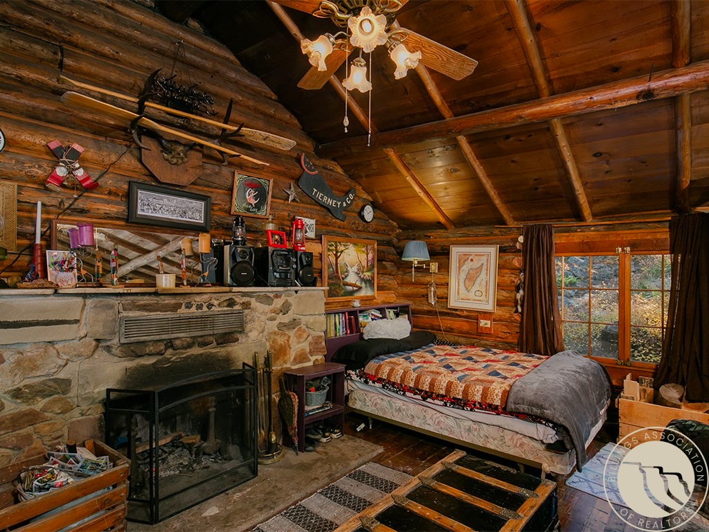 Log-cabin bedroom with a stone fireplace, patchwork bedspread, timber ceiling, and window overlooking autumn foliage.