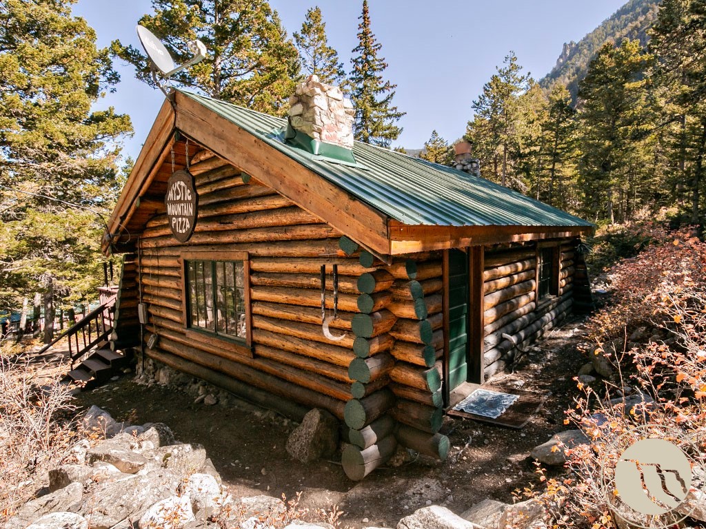 Log cabin with a green metal roof and stone chimney among trees on a rocky hillside.