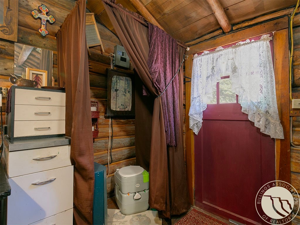 Rustic log bathroom with a red door, lace curtain, brown drapes, and portable toilet.