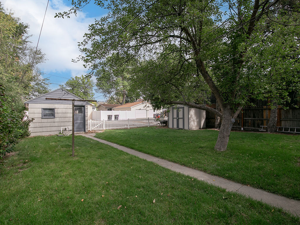 Backyard with a concrete walkway, mature shade tree, two sheds, and a white picket fence.