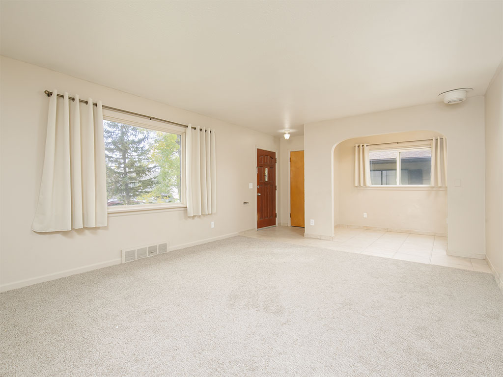Empty carpeted living room with curtained windows and an arched opening to a tiled area.
