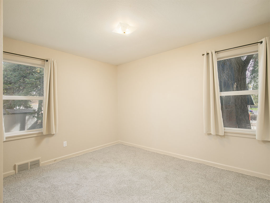 Empty bedroom with beige walls, carpet, and two curtained windows.