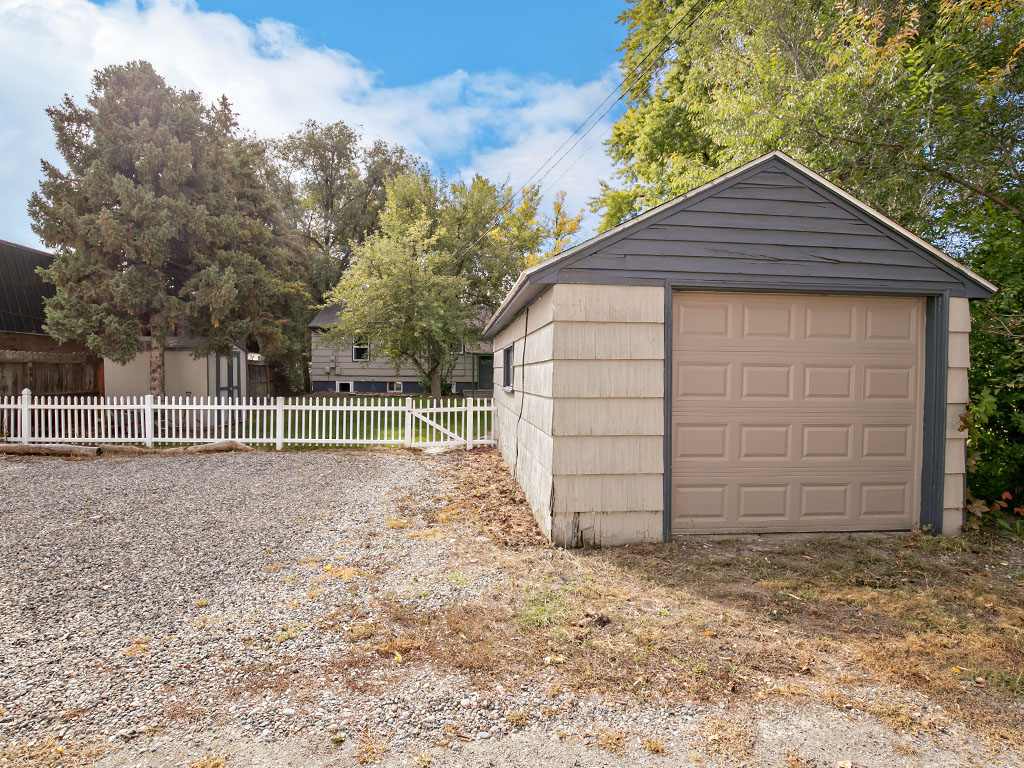 Detached garage with a tan door beside a gravel driveway and white picket fence.