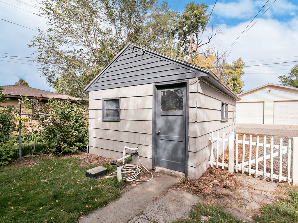Gray-sided outbuilding with a dark door beside a white picket fence.