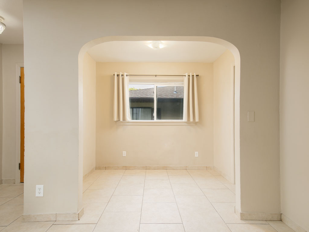 Vacant dining area with a wide archway, pale tile floor, and curtained window.