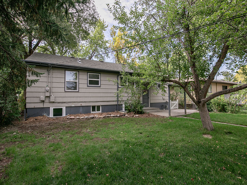 Gray-sided single-story house framed by mature trees, with a green lawn and covered entry.