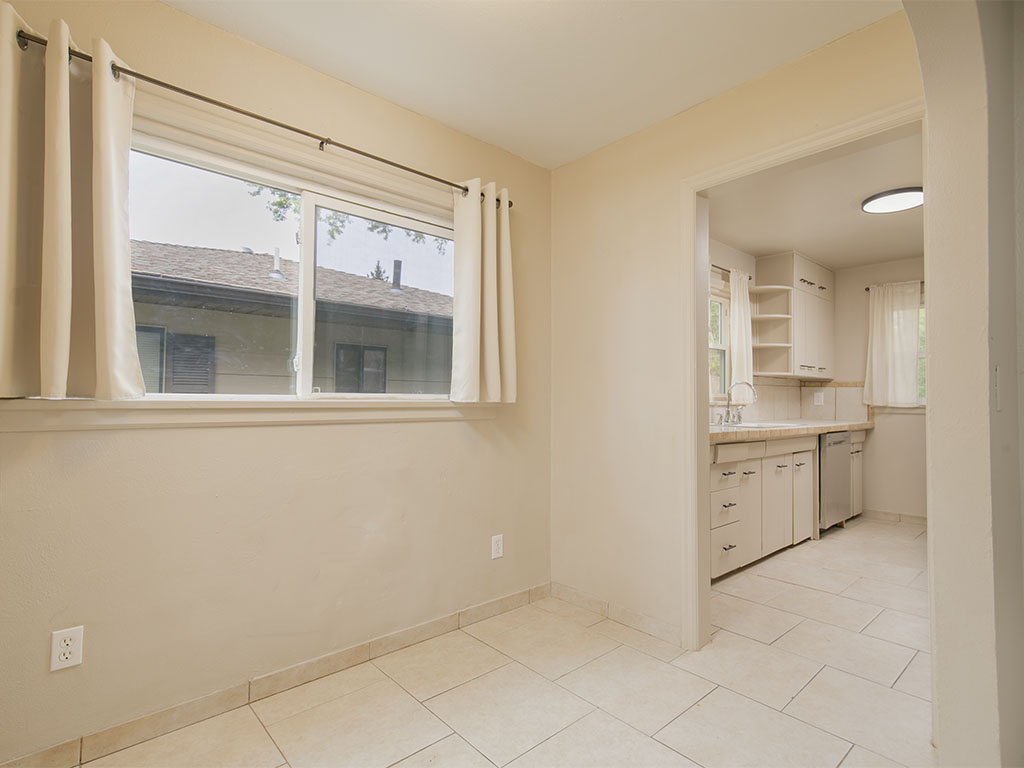 Empty tiled dining area with a wide curtained window and an adjoining white-cabinet kitchen.
