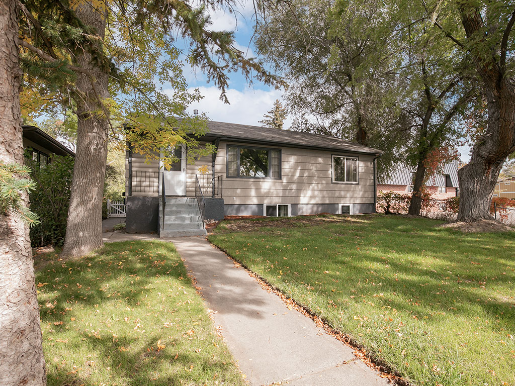Single-story beige-sided home framed by mature trees, with front steps and a walkway across the lawn.