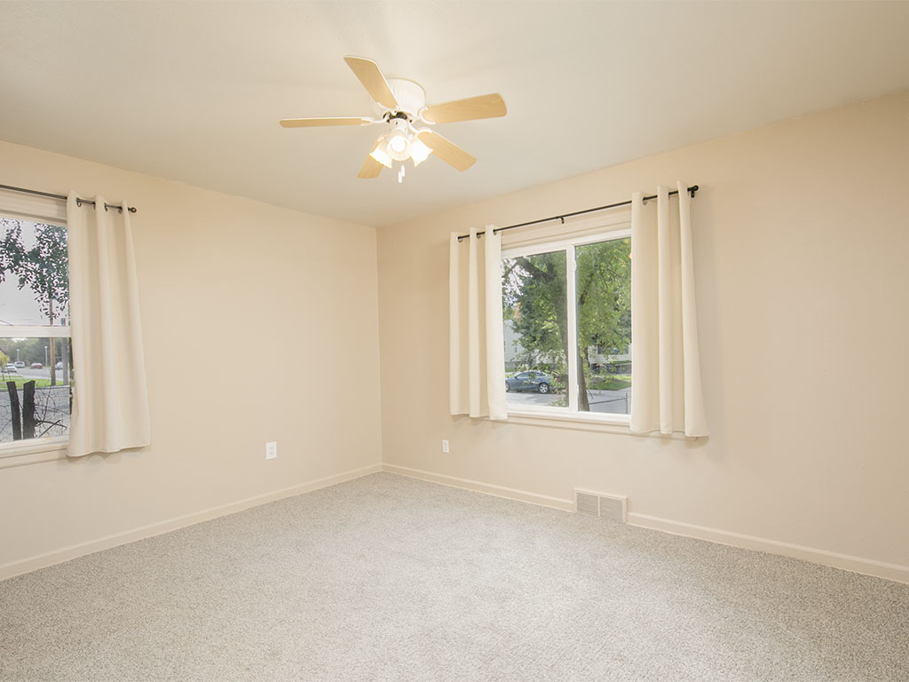 Empty bedroom with light carpet, two curtained windows, and a ceiling fan.