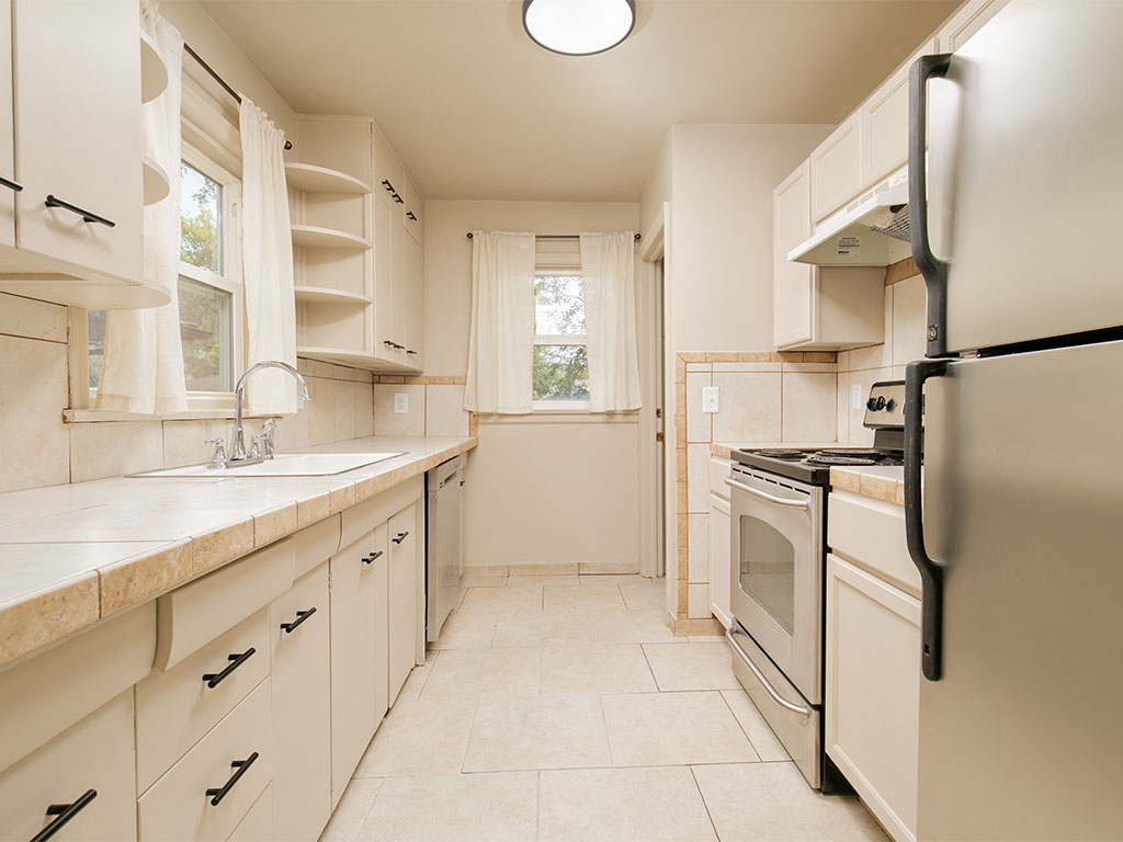 Bright narrow kitchen with white cabinets, tiled counters, and a window above the sink.