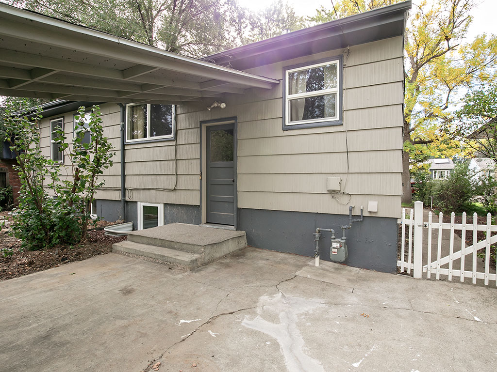 House side entrance beneath a covered carport, with a concrete driveway and white picket gate.