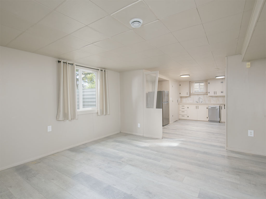 Vacant living area with pale plank flooring, opening to a white kitchen with stainless steel appliances.