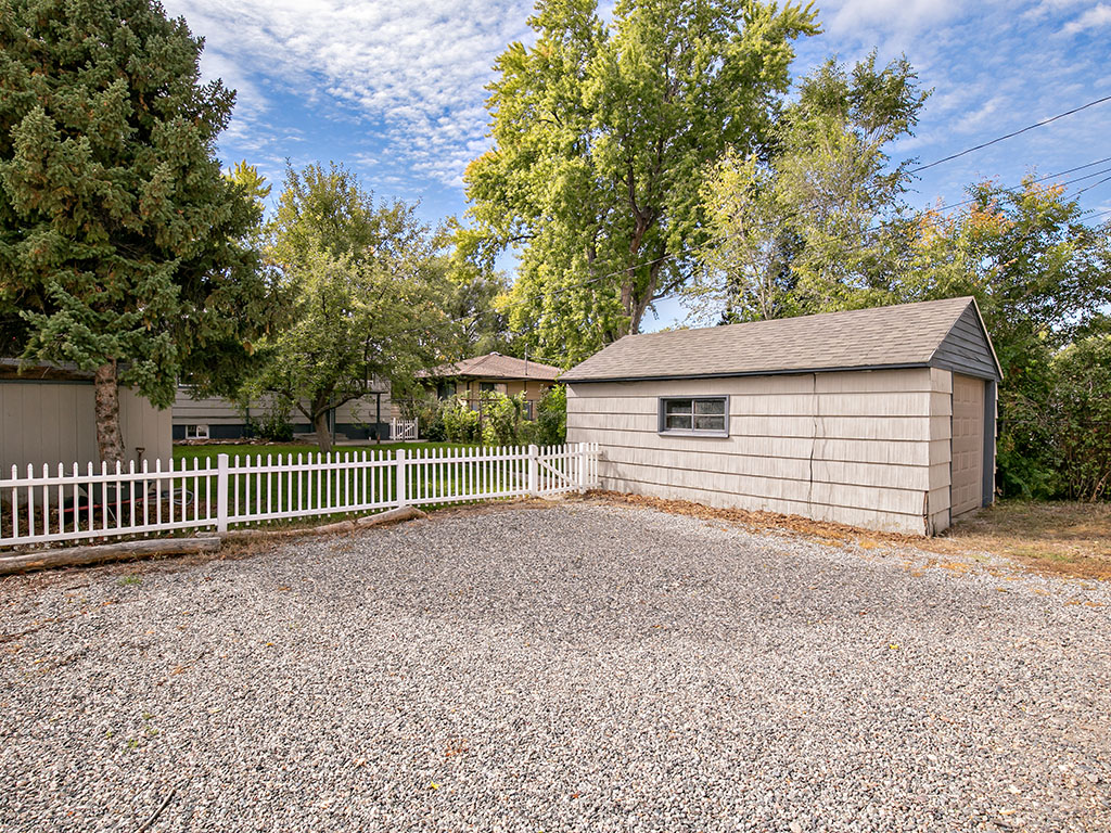 Detached beige garage beside a gravel driveway and white picket fence, framed by mature trees.