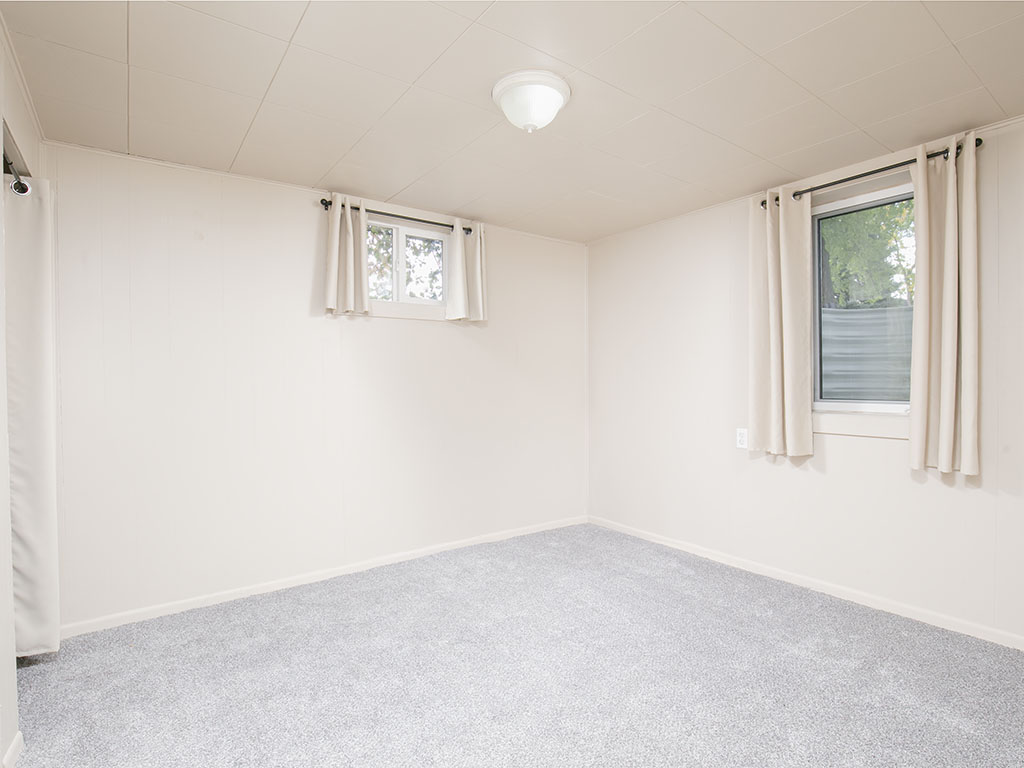 Empty carpeted room with cream walls, two curtained windows, and a small high-set window.