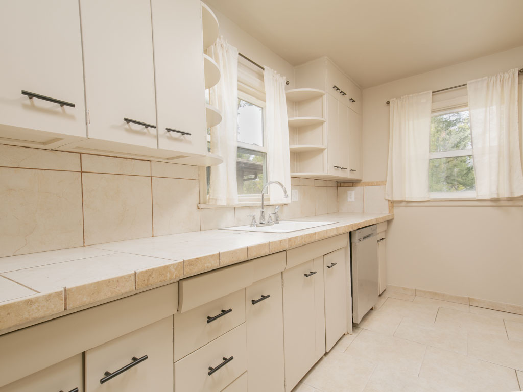Kitchen with white cabinetry, tiled counters, a sink beneath a window, and a dishwasher.